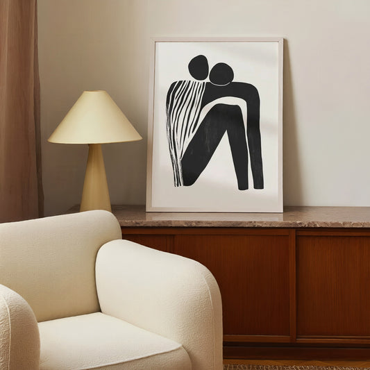 Framed abstract artwork on a wall above a wooden cabinet with a lamp and beige armchair.