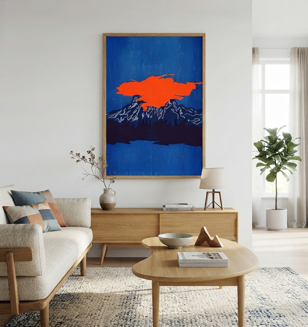 A framed abstract landscape art print featuring a bright orange cloud over a blue mountain and lake, displayed on a white wall in a modern, sunlit living room with a wooden console, sofa, and coffee table.