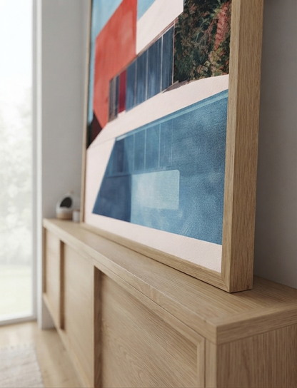 A large framed Bauhaus style swimming pool architectural print with red and blue geometric house with blue, red, and white geometric shapes leans against a light wood console table in a modern room.