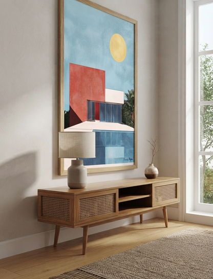 A large framed Bauhaus style swimming pool architectural print with colourful modern architecture hangs above a mid-century modern wooden console table in a bright, modern room with a window, lamp, and rug.
