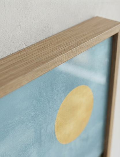 Close-up of a framed Bauhaus style swimming pool architectural print with abstract house illustration with a light wood frame, textured light blue background, and a large gold circle.