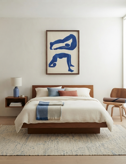 A modern bedroom scene featuring a low wooden platform bed with cream bedding, a blue throw blanket, a matching blue nightstand lamp, and a framed Matisse-Inspired Blue Nude Print for Living Room by PrintHaus on the wall above the headboard.