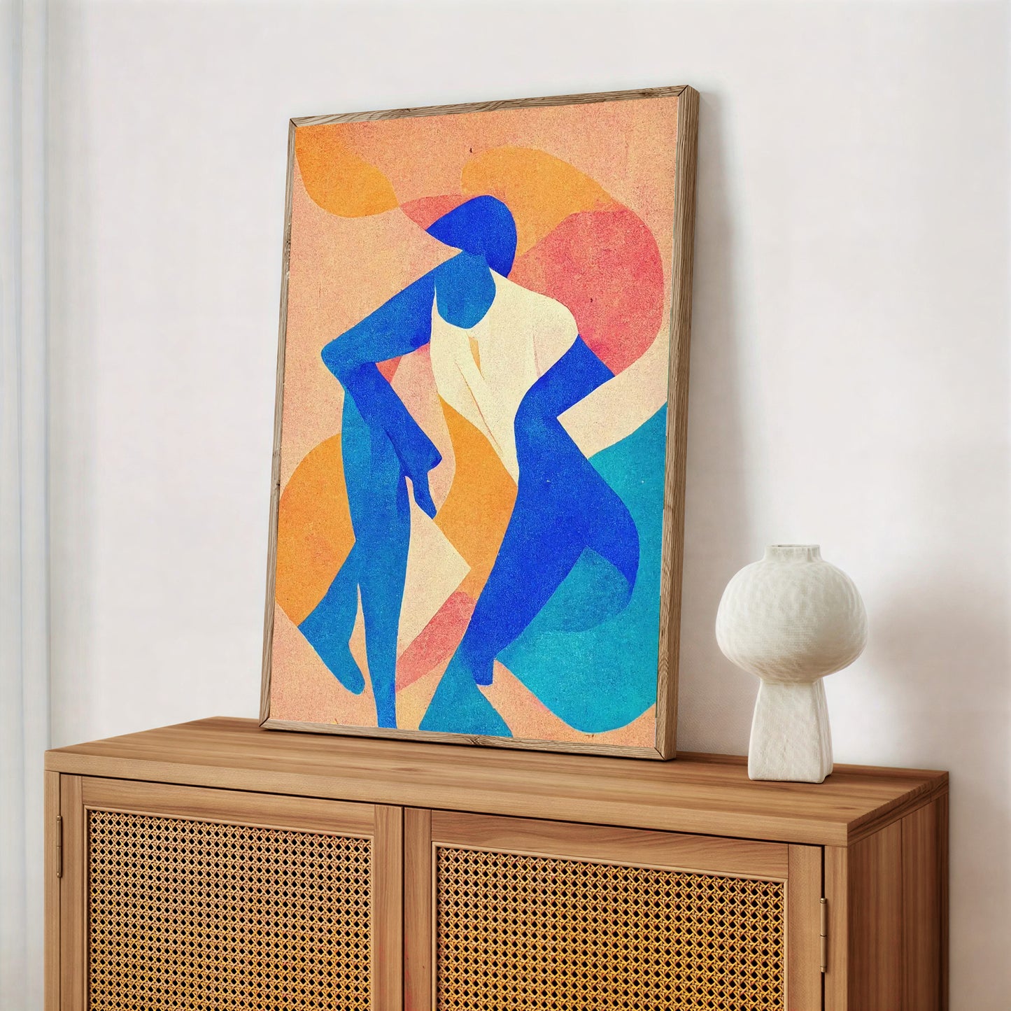 Framed abstract artwork of two blue figures on a wooden cabinet with a white vase.