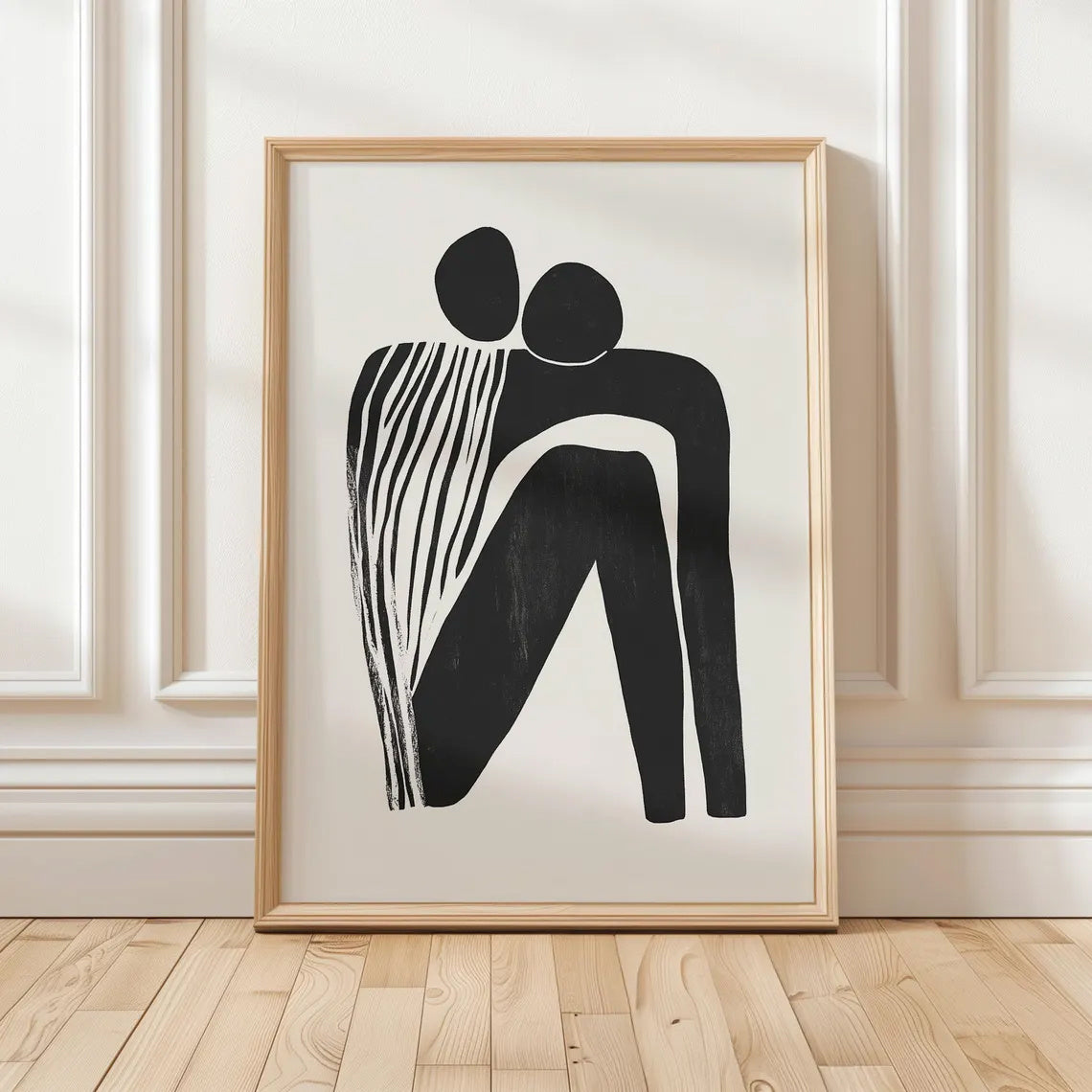 Framed abstract artwork of two black figures on a white background, displayed on a wooden floor with a white paneled wall.