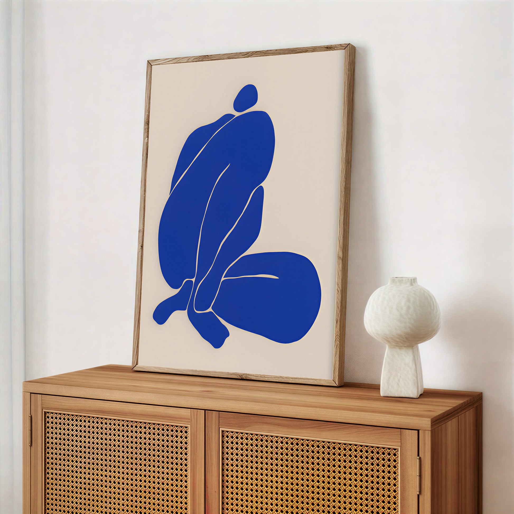 Framed abstract artwork with a blue figure on a beige background, displayed on a wooden cabinet.