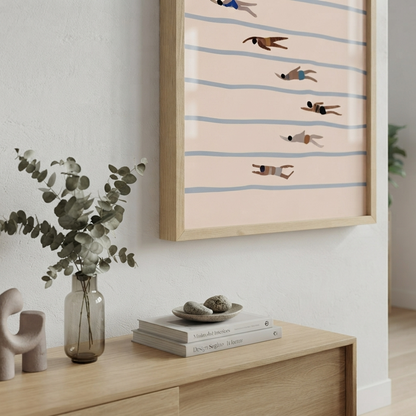 Framed Minimalist Swimming Pool Art Print inspired by mid-century modern swimming pool art,  featuring a minimalist illustration of people floating on horizontal blue stripes against a pale pink background, displayed in a light wood frame on a white wall above a wooden console table with books and a vase.