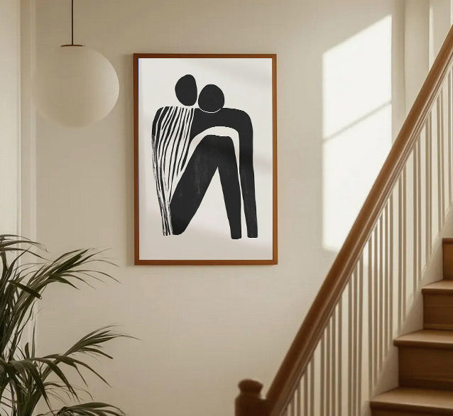Framed abstract artwork of two figures on a wall in a home setting with a staircase.