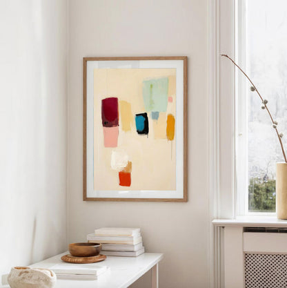 Abstract art print in a wooden frame on a wall with a window and books in the foreground.