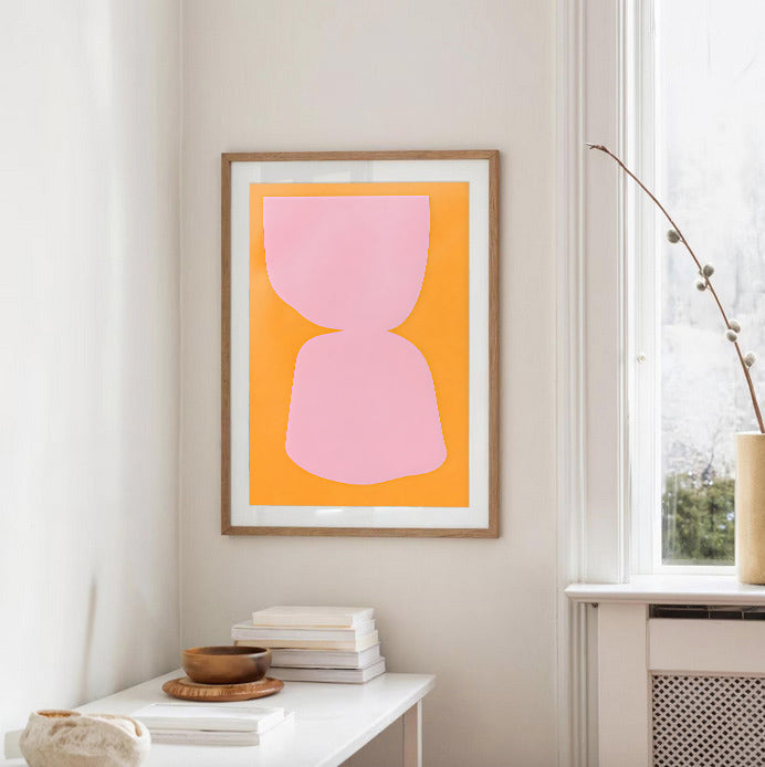 Framed abstract art with a pink shape on an orange background in a room with a window and books.