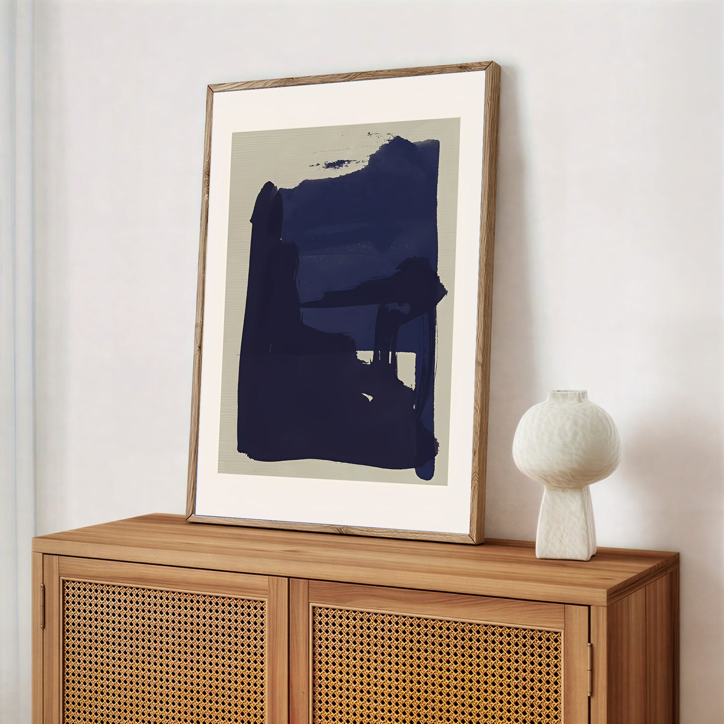 Framed abstract art piece on a wooden cabinet with a white vase.