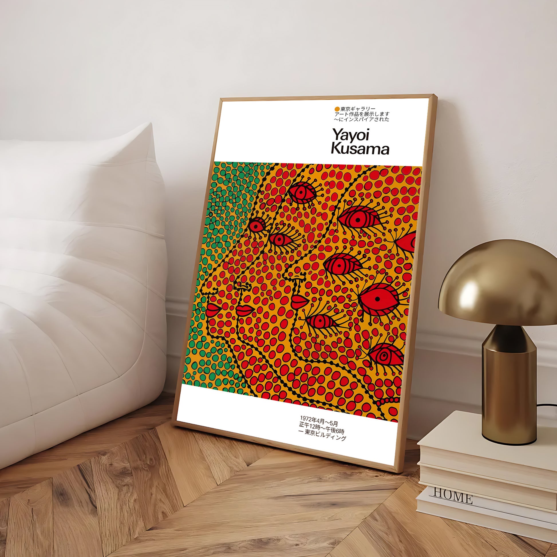 Framed Yayoi Kusama art poster leaning against a wall beside a white sofa and a gold lamp, featuring a colorful dotted pattern with stylized faces and red eyes