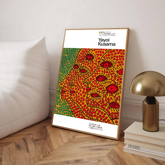 Framed Yayoi Kusama art poster leaning against a wall beside a white sofa and a gold lamp, featuring a colorful dotted pattern with stylized faces and red eyes