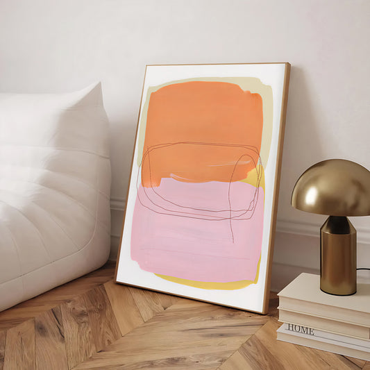 Framed abstract art with orange and pink colors on a wooden floor next to a white pillow and gold lamp.