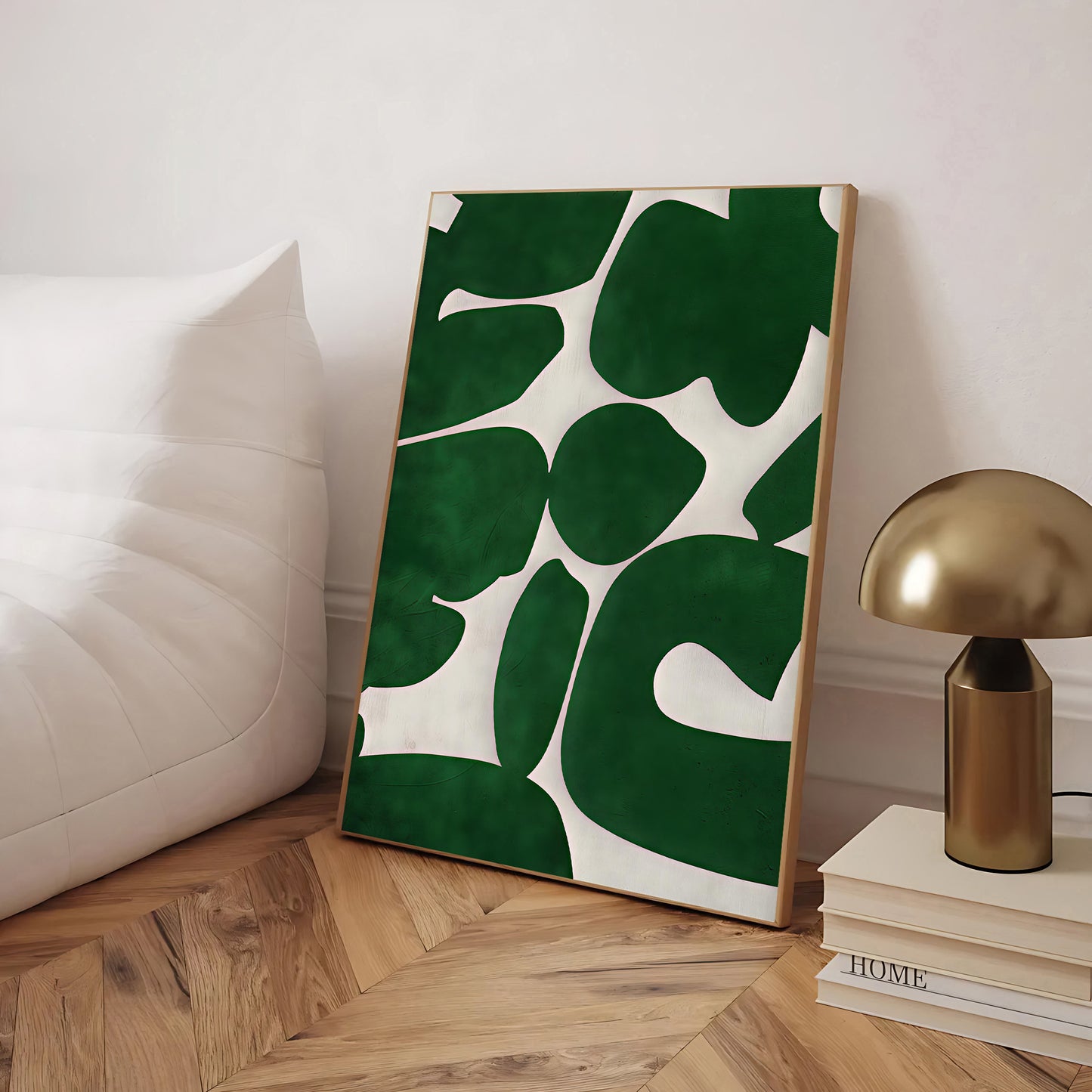 Framed artwork with green leaf pattern on a wooden floor next to a gold lamp and books.