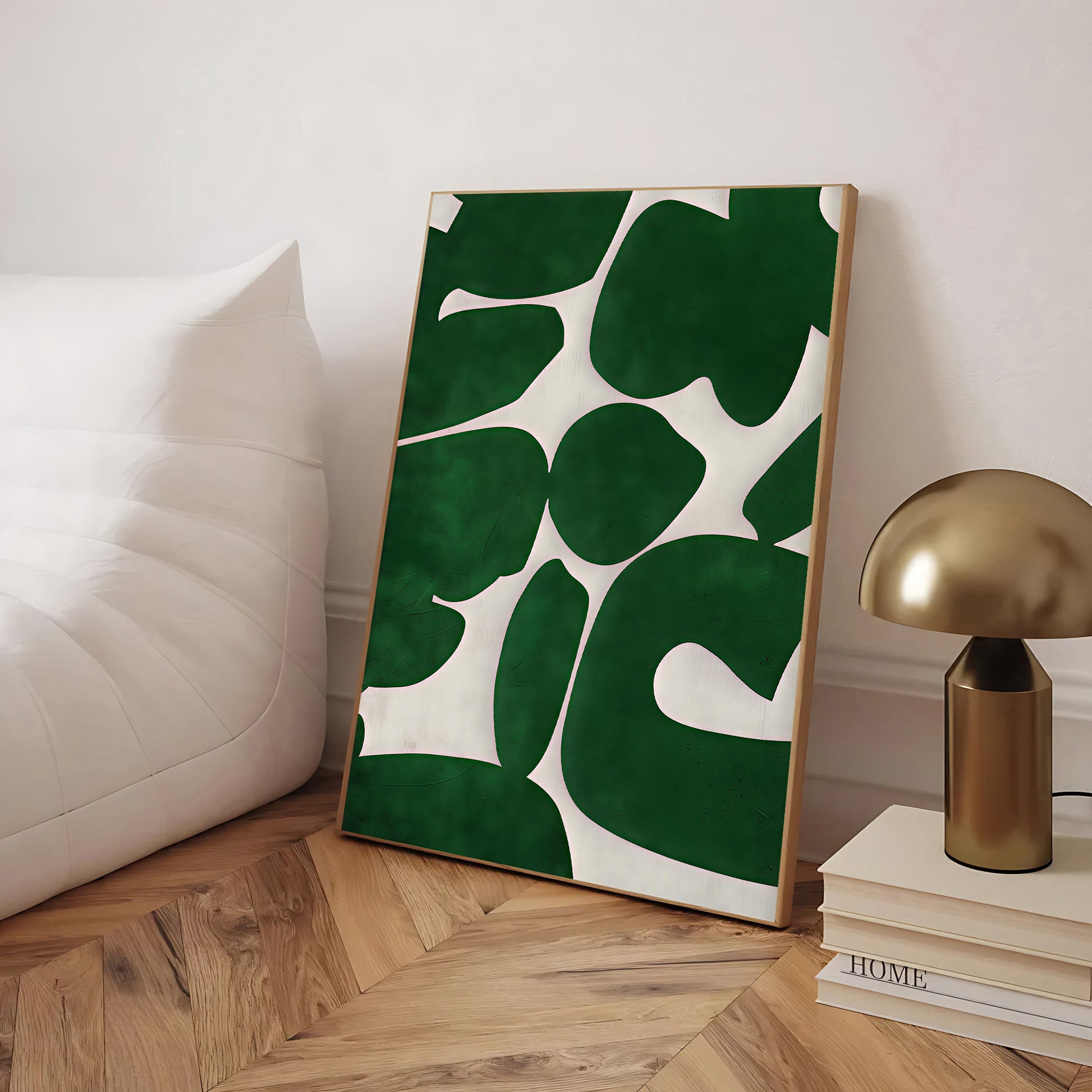 Framed artwork with green leaf pattern on a wooden floor next to a gold lamp and books.