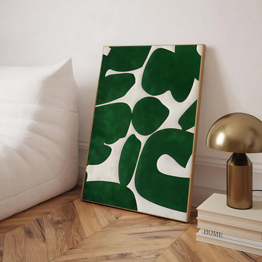 Framed artwork with green leaf pattern on a wooden floor next to a gold lamp and books.