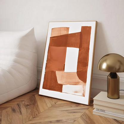 Abstract art print in a wooden frame on a wooden floor with a lamp and books in the background.