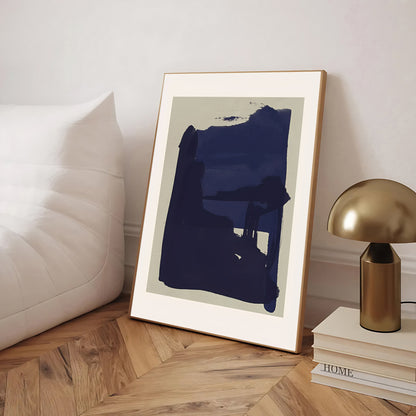 Framed abstract art piece on a wooden floor with a lamp and books in the background.