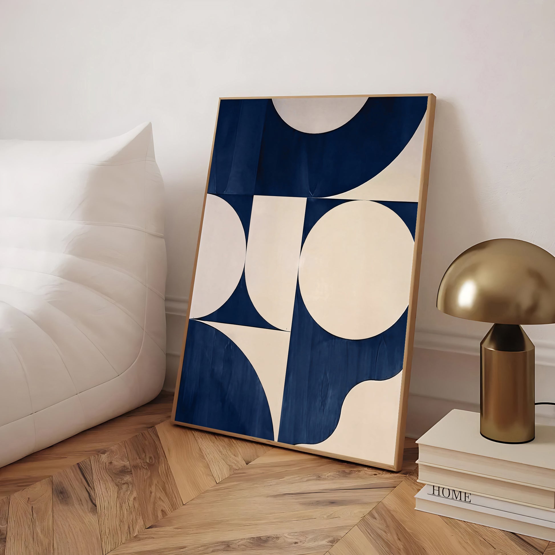 Framed abstract art piece with blue and white circles on a wooden floor, next to a gold lamp and books.
