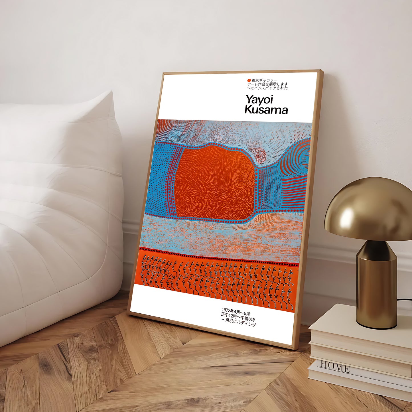 Framed artwork by Yayoi Kusama on a wooden floor with a lamp and books in the background.