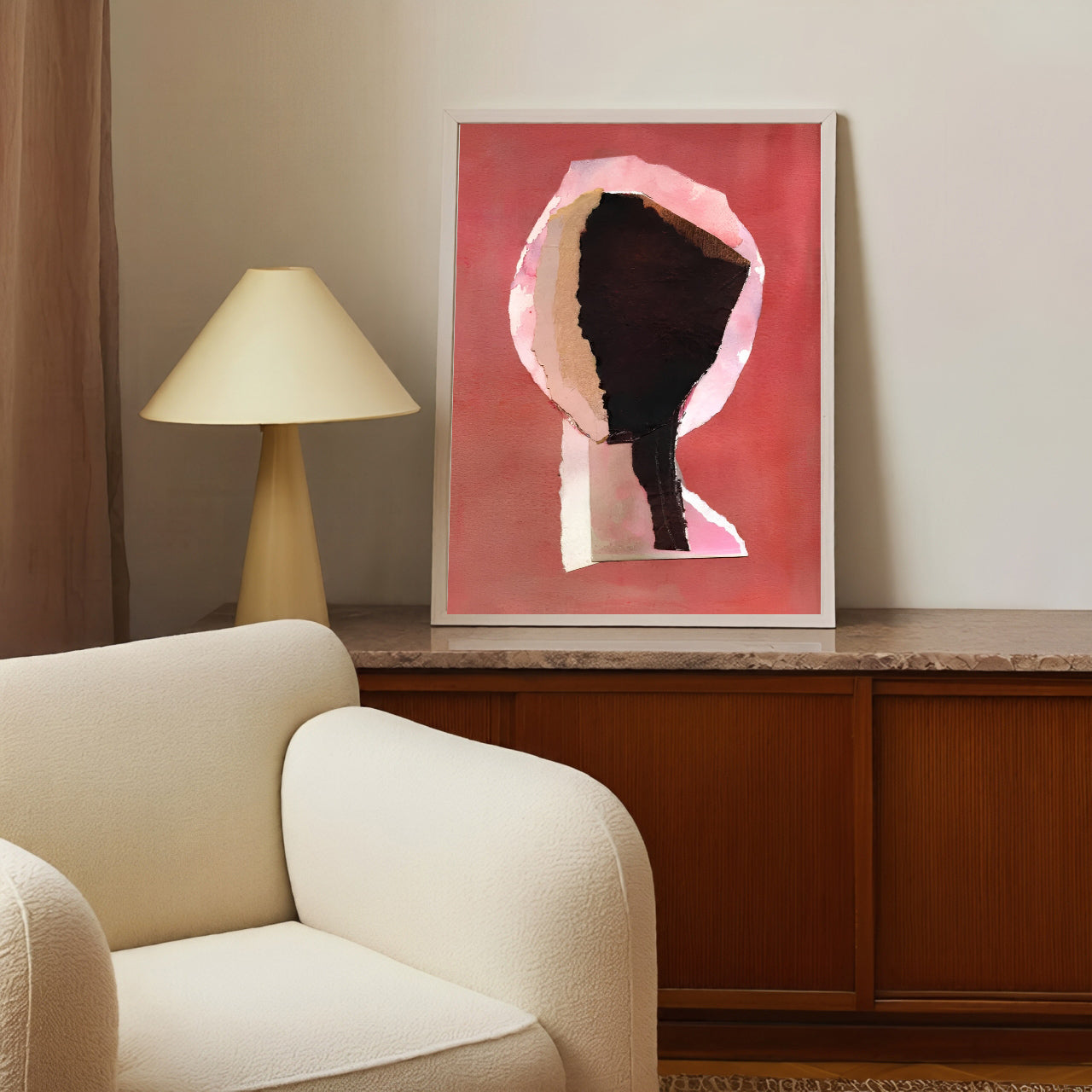 Framed abstract art on a wall above a wooden cabinet with a lamp and beige armchair.