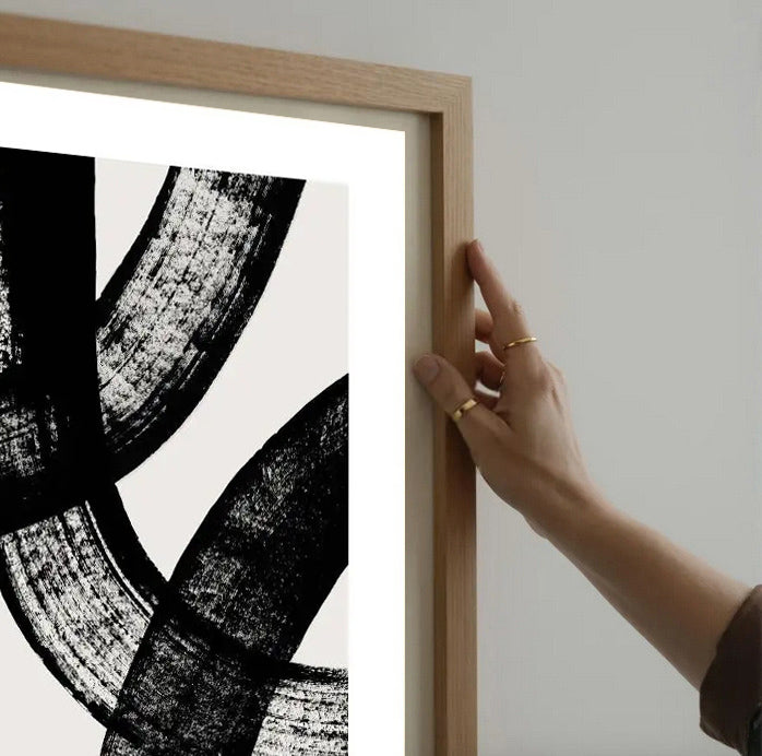 Person holding a wooden-framed abstract black and white artwork on a light gray wall.